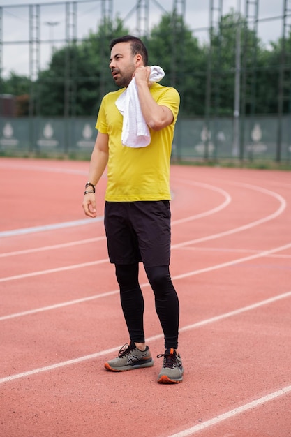 Exhausted athlete wipes his neck with a soft towel while standing on the running track