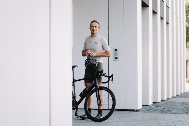 Exhausted caucasian cyclist standing near black bike with safety helmet in hands Male athlete taking break during morning exercise at urban area