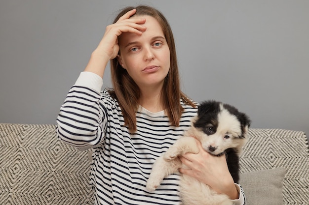 Exhausted Caucasian woman wearing striped shirt sitting on sofa with small dog being tire playing wi