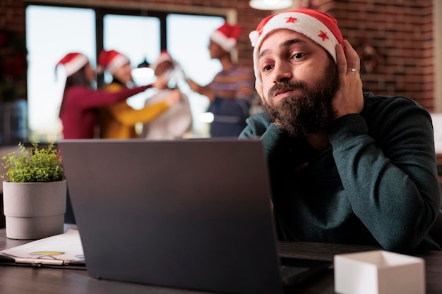 Exhausted distrubed person trying to work in office, feeling frustrated and overwhelmed because of noisy colleagues in workplace decorated with christmas ornaments. Dissatisfied unhappy worker.