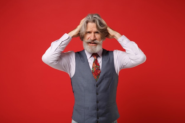 Exhausted elderly gray-haired mustache bearded man in classic shirt vest colorful tie isolated on red background, studio portrait. People lifestyle concept. Mock up copy space. Putting hands on head.