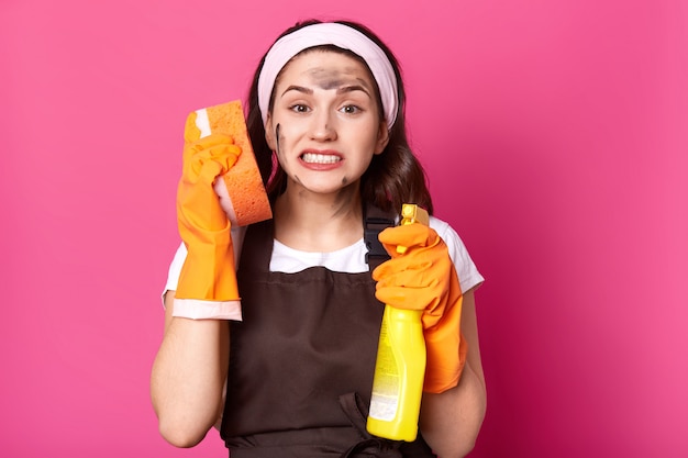 Exhausted emotional cleaner standing isolated over pink in studio, holding washcloth near ear and detergent