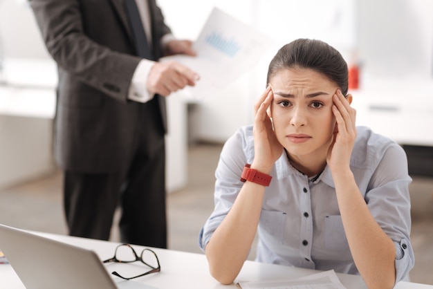 Exhausted girl sitting at her workplace leaning her head on the hands pressing lips while looking at front