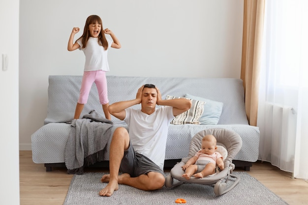 Exhausted handsome brunette father sitting on floor near sofa with smaller daughter in bouncer, wearing white t shirt and jeans short, man covering ears from screaming elder daughter.