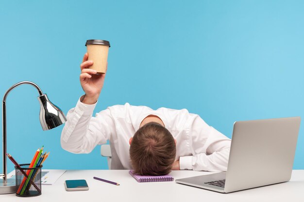 Photo exhausted male employee feeling fatigue lying on table and raising coffee cup lack of energy in morning office tired of stress problems sleeping at workplace overwork concept indoor studio shot