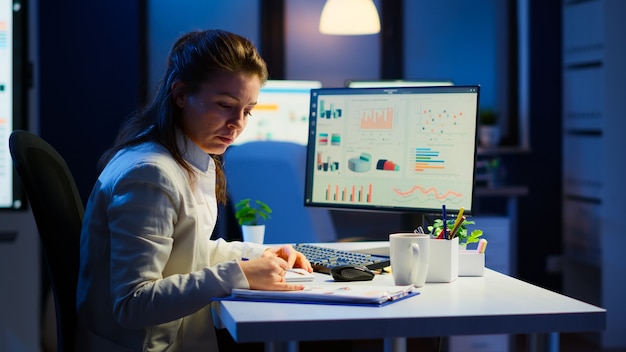 Exhausted manager trying to finish business project respecting deadline working at night in front of computer taking notes writing on notebook. Tired employee overtime sitting at desk in workplace