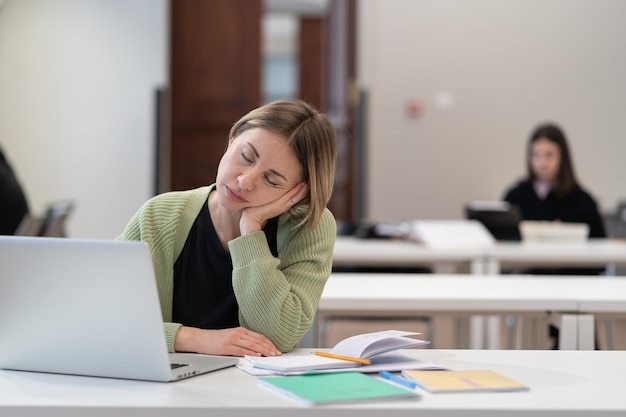 Exhausted mature female student falling asleep during online lesson studying on laptop in classroom