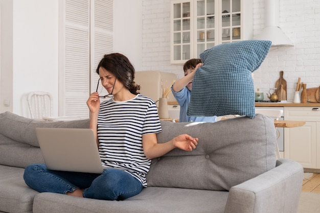 Exhausted mother sitting on couch at home work on laptop, child distracts, attacking mom with pillow