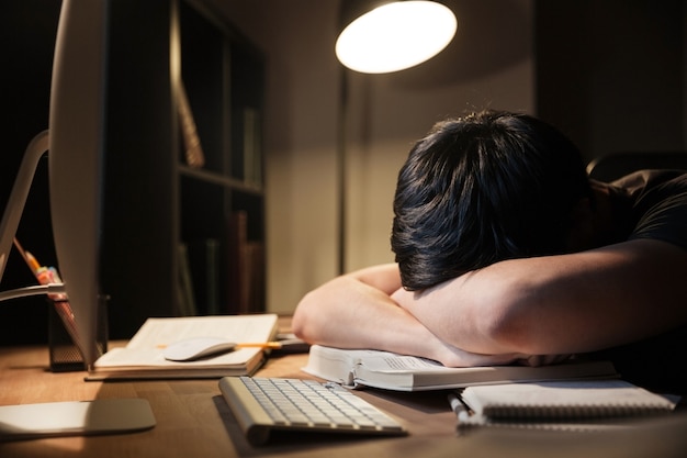 Exhausted overworked young man studying and sleeping on the table in dark room at home