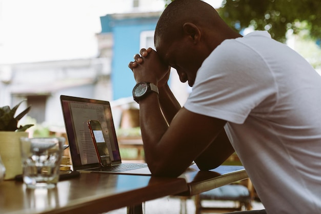 Exhausted stressed dark skin man leaning his head on joint hands near laptop and smartphone Online conference in cafe