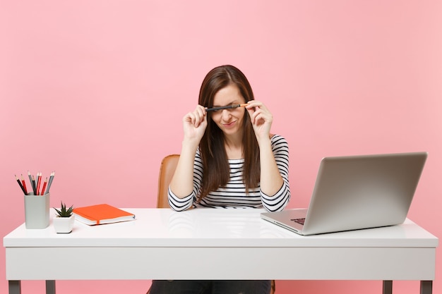 Exhausted tired woman having problem holding pencil near face sit, work at white desk with contemporary pc laptop isolated on pastel pink background. Achievement business career concept. Copy space.