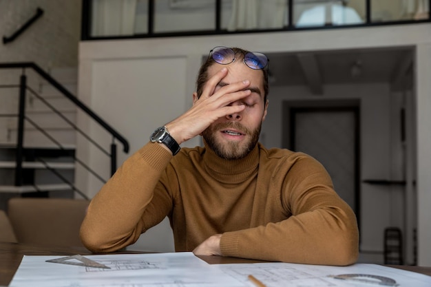 Exhausted tired young bearded young engineer architect working on new design of house creating blueprint being overworked