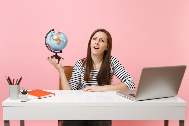 Exhausted upset woman holding globe having problems with vacation planning while sit, work at office with pc laptop isolated on pastel pink background. Achievement business career concept. Copy space.