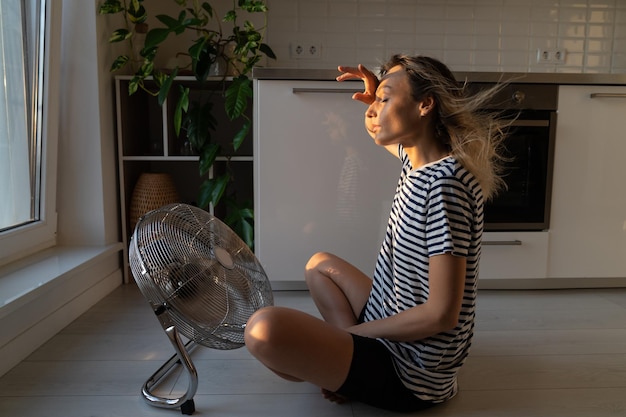 Exhausted woman refreshing sit near big indoor ventilator blowing cooling fresh air at home on floor