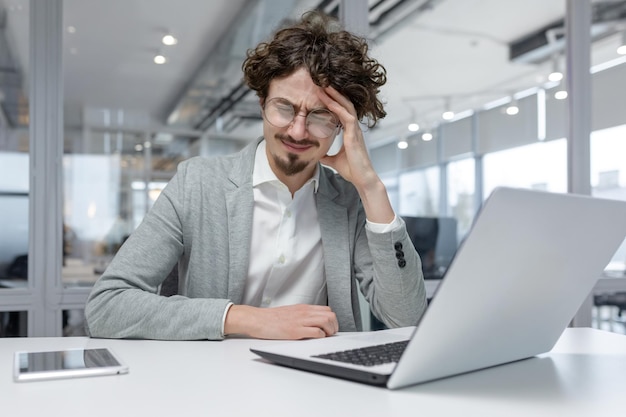 Exhausted young businessman at his desk in a bright office rubbing his temples feeling stressed