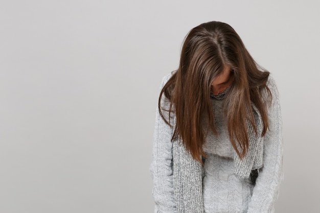 Exhausted young woman in gray sweater, scarf with lowered head isolated on grey background, studio portrait. Healthy fashion lifestyle, people sincere emotions cold season concept. Mock up copy space.