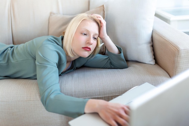 Exhausted young woman lying on sofa using laptop too tired or bored of online work at home free space Workaholism chronic fatigue overworking on remote job concept