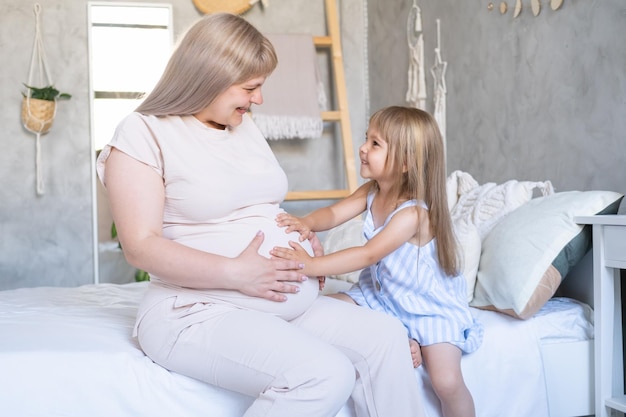 Expectant mother sitting on bed with little baby daughter stroking belly