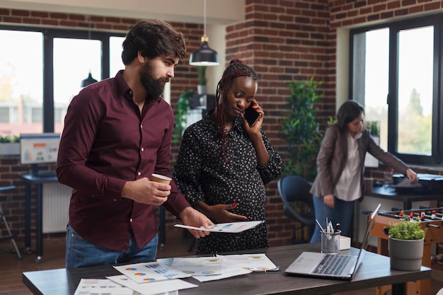 Expectant mother talking on phone in financial agency office workspace while department colleague reading important paperwork. Company worker reviewing project marketing strategy ideas.