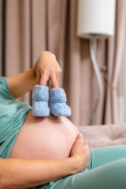 Expecting mother sitting on the sofa and holding her fingers in tiny blue booties Socks for a little boy on a big pregnant tummy Blurred curtains and lampshade at the backdrop