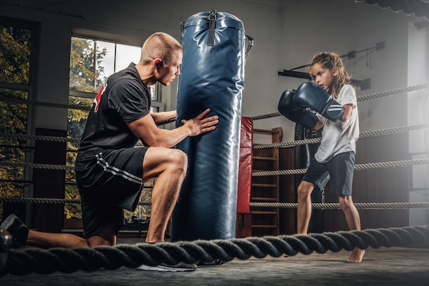 Experienced boxer trainer is training new little girl boxer for special competitions using punching bag.