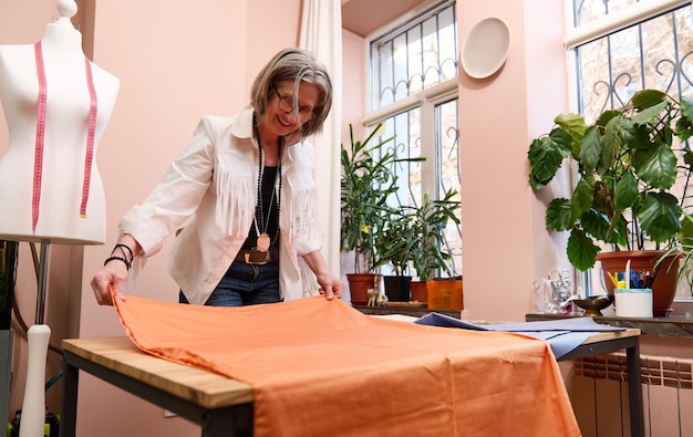 Experienced elderly female fashion designer tailor laying out an orange colored fabric on a wooden table creating new garment in tailoring atelier