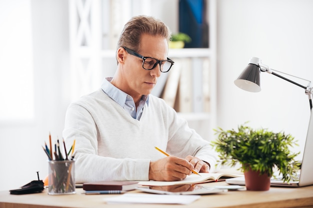 Experienced expert at work. Confident mature man writing something in his notebook while sitting at his working place