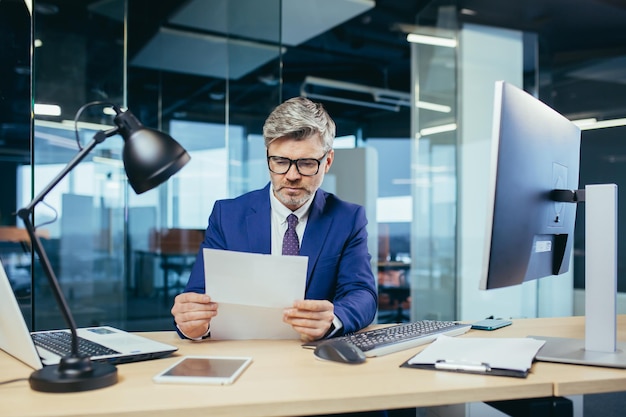 Experienced grayhaired businessman working at a computer paperwork in the office reading a letter from the bank