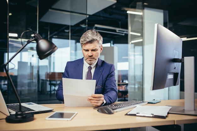 Experienced grayhaired businessman working at a computer paperwork in the office reading a letter from the bank