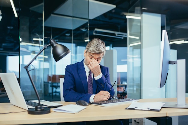 Photo an experienced grayhaired businessman works at the computer until late tired has a severe headache stress and depression