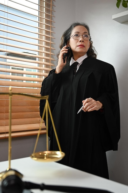 Experienced mature female lawyer standing in her office and talking on mobile phone with her clients for providing law consultation