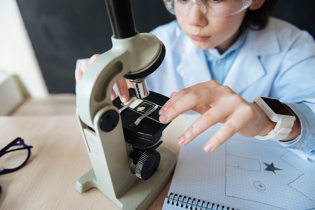 Exploring evolution of microbiology. Skillful gifted capable teenager sitting in the laboratory and enjoying science lesson while makig notes and using microscope