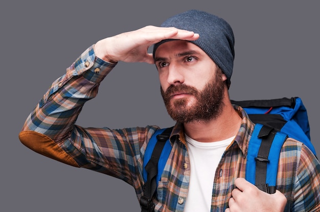 Exploring new places. Handsome young bearded man carrying backpack and looking at view while holding hand upon his eyes and standing against grey background