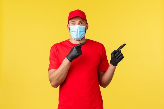 Express delivery during pandemic, covid-19, safe shipping, online shopping concept. Excited courier in red uniform, merical mask and gloves, pointing right, tell big news