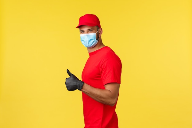 Express delivery during pandemic, covid-19, safe shipping, online shopping concept. Friendly male courier in red uniform and medical mask, provide safe contactless package deliver, thumb-up