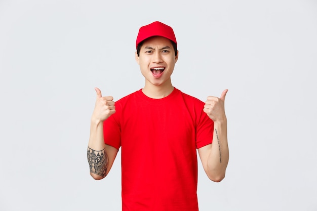 Express delivery, shipping and logistics concept. Upbeat handsome asian courier in red t-shirt and cap, show thumbs-up, encourage keep up good work, being the best, chanting in celebration