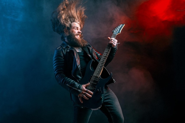 Expression rock guitar player with long hair and beard plays on the smoke background studio shot