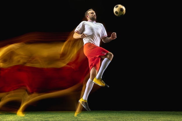 Expression. Young caucasian male football or soccer player in sportwear and boots kicking ball for the goal in mixed light on dark wall. Concept of healthy lifestyle, professional sport, hobby.