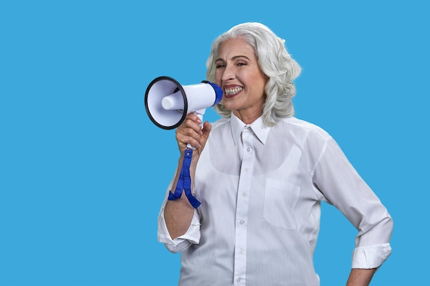 Expressive senior woman in white blouse talking into megaphone mature lady with loudspeaker on blue