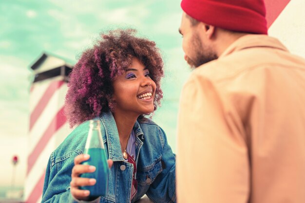 Expressive talk. Excited young girl sitting with a bottle of blue drink in her hand and looking at her boyfriend while smiling to him