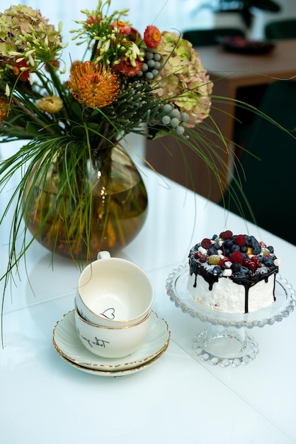 An exquisite berry cake on a glass stand on a table with a vase of flowers and a tea set in a modern designer interior. Soft selective focus, art noise