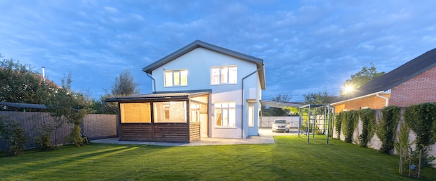 Exterior view of modern white house with courtyard, patio area, green grass lawn, garden and a car in the evening