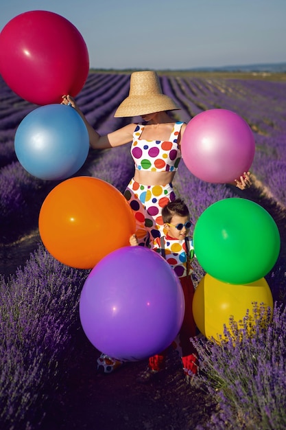 Photo extravagant woman mother in a white suit with multicolored polka dots ,and hat stands in a lavender field with her son in a multicolored clown costume