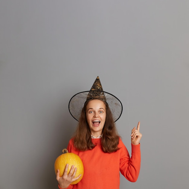 Extremely amazed winsome woman dressed in witch hat holding orange pumpkin isolated on gray background pointing finger up at copy space for advertisement or promotional text