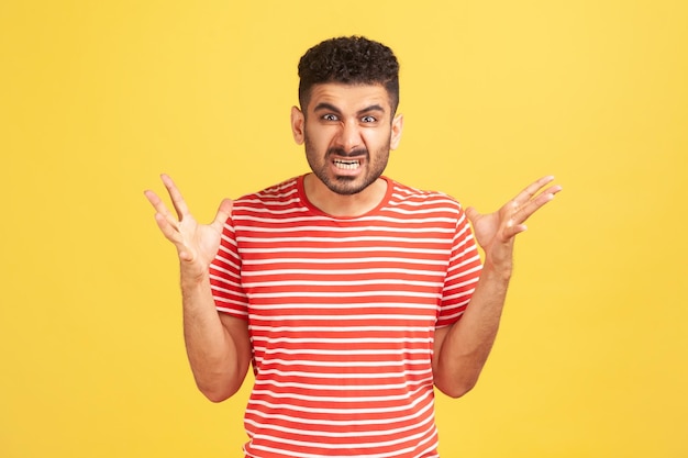 Extremely angry furious bearded man in striped t-shirt clenching teeth and spreading hands, looking at camera with nervous expression, dissatisfaction. Indoor studio shot isolated on yellow background