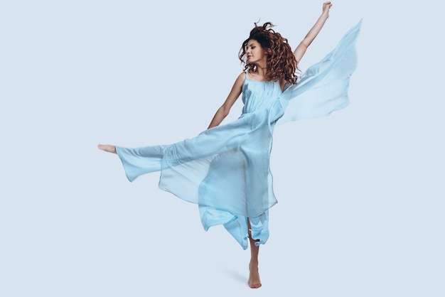 Extremely beautiful. Full length studio shot of attractive young woman in elegant dress gesturing and smiling while jumping against grey background