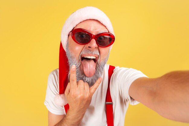 Photo extremely excited crazy man in santa claus hat and sunglasses fooling faces looking at camera, elderly blogger having fun showing rock gesture. indoor studio shot isolated on yellow background