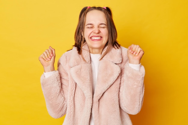 Extremely happy brown haired girl wearing pink fur coat with ponytails isolated over yellow background does winner gesture clenches fists rejoices positive news glad to achieve goal