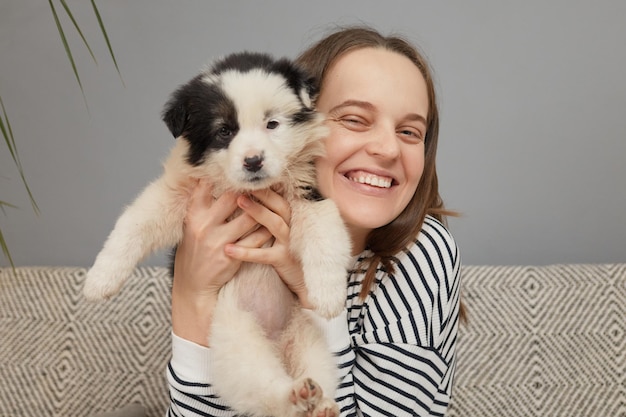 Extremely happy brown haired woman dressed in striped shirt embracing small puppy looking at camera