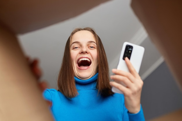 Extremely happy Caucasian woman unpacking box and looking in cardboard box with happy smile looks joyful holding smart phone in hands received long awaited parcel view from the inside of package